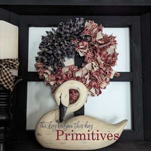 May include: A white wooden swan figurine with the text "The day before this day. Primitives" on the base. The swan is decorated with a red, white and blue rag wreath.