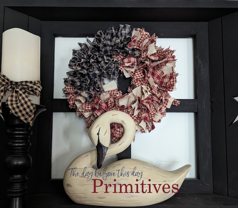 May include: A white wooden swan figurine with the text "The day before this day. Primitives" on the base. The swan is decorated with a red, white and blue rag wreath.