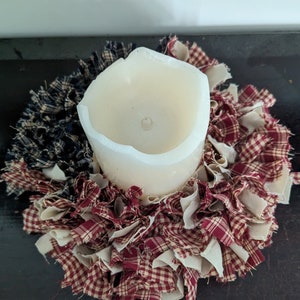 May include: A white pillar candle sits in the centre of a red and white gingham fabric wreath. The wreath is made of strips of fabric that are frayed and tied together.