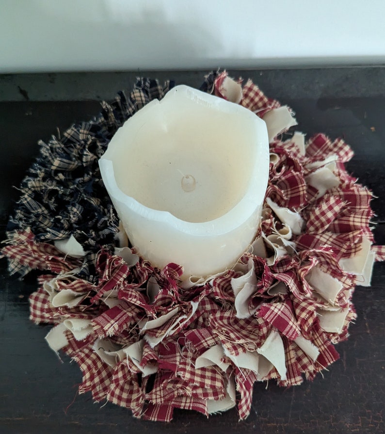 May include: A white pillar candle sits in the centre of a red and white gingham fabric wreath. The wreath is made of strips of fabric that are frayed and tied together.