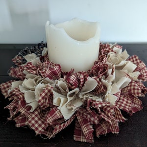 May include: A white candle sits in the centre of a red and white gingham fabric wreath. The wreath is made of fabric scraps and is a rustic, farmhouse style decoration.