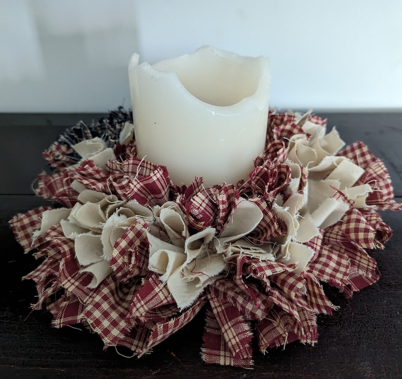 May include: A white candle sits in the centre of a red and white gingham fabric wreath. The wreath is made of fabric scraps and is a rustic, farmhouse style decoration.
