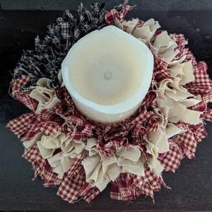 May include: A white pillar candle sits in the centre of a red, white and blue fabric wreath. The wreath is made of fabric scraps and is a rustic, handmade design.
