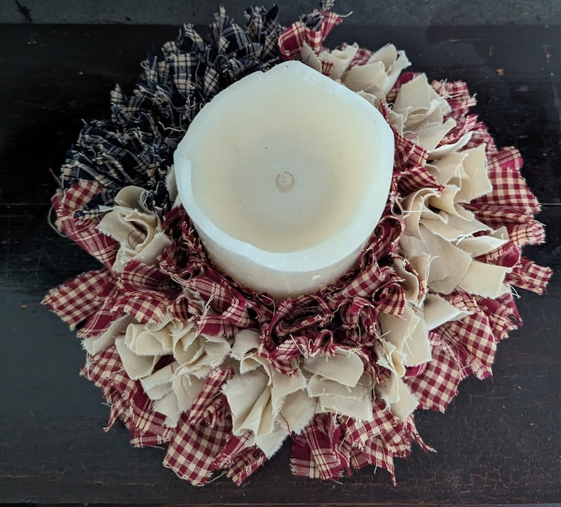 May include: A white pillar candle sits in the centre of a red, white and blue fabric wreath. The wreath is made of fabric scraps and is a rustic, handmade design.