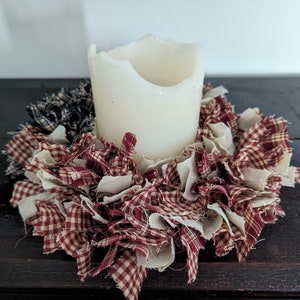 May include: A white candle sits in the centre of a red and white gingham fabric wreath. The wreath is made of strips of fabric that have been frayed and tied together.