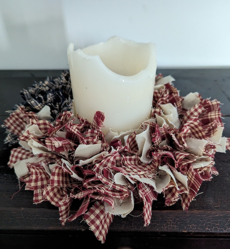 May include: A white candle sits in the centre of a red and white gingham fabric wreath. The wreath is made of strips of fabric that have been frayed and tied together.