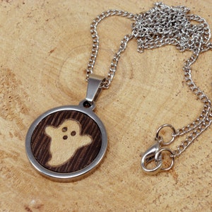 May include: A silver chain necklace with a round pendant featuring a wood-burned ghost design. The pendant is made of dark wood with a light wood ghost design.