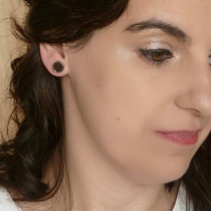 May include: A close-up of a woman's face with dark brown hair. She is wearing black round earrings. Her makeup includes eyeshadow and eyeliner.