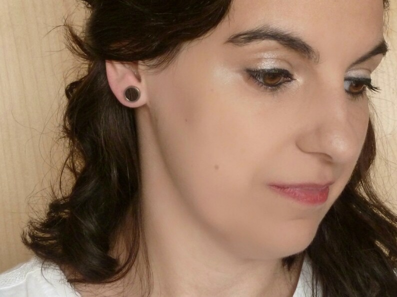 May include: A close-up of a woman's face with dark brown hair. She is wearing black round earrings. Her makeup includes eyeshadow and eyeliner.