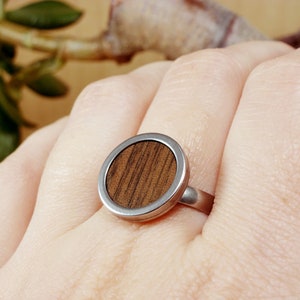 May include: A silver ring with a round wooden inlay. The wood has a dark brown color and a striped pattern.