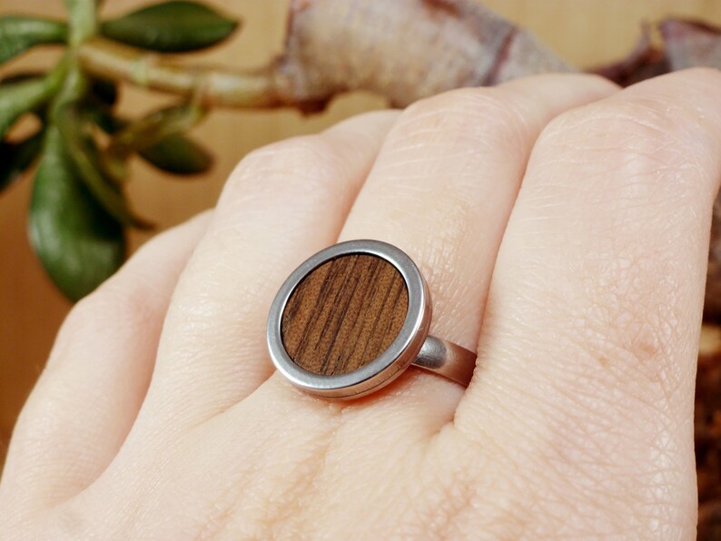 May include: A silver ring with a round wooden inlay. The wood has a dark brown color and a striped pattern.