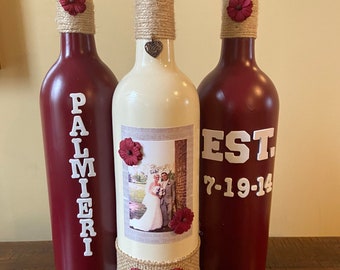 May include: Three wine bottles decorated for a wedding. The centre bottle is white with a photo of a bride and groom, burlap ribbon, and red flowers. The bottles on either side are burgundy with the words "Palmieri" and "Est. 7-19-14" written on them.
