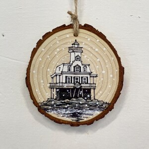 May include: A round, wooden Christmas ornament with a hand-painted lighthouse scene. The black and white lighthouse sits on a rocky base, with falling snow. The ornament is made from a cross-section of a tree branch and has a natural wood grain.