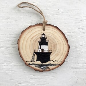 May include: A round wooden ornament with a painted black and white lighthouse design. The ornament is made from a cross-section of a tree branch, with visible wood grain and a natural edge. A twine loop is attached for hanging.