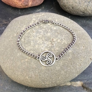 May include: A silver chain bracelet with a silver Celtic knot charm. The bracelet is on a gray stone background.