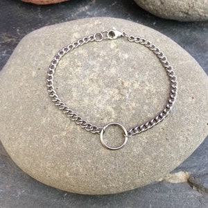 May include: A silver chain bracelet with a circular charm. The bracelet is on a grey stone background.