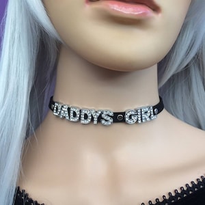 May include: Black choker necklace with silver metal letters that spell "DADDY'S GIRL" and are decorated with clear rhinestones.