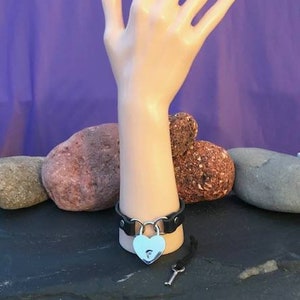 May include: Black leather bracelet with a heart-shaped padlock and a silver key. The bracelet is on a mannequin hand.