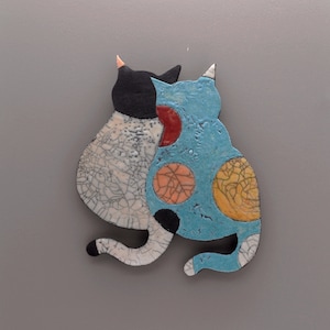 Puede incluir: Una obra de arte en cerámica que presenta dos figuras de gatos estilizadas. Un gato es blanco y negro, el otro es azul con manchas naranjas y amarillas. Los gatos están cerca, sugiriendo afecto. La obra tiene una superficie texturizada.