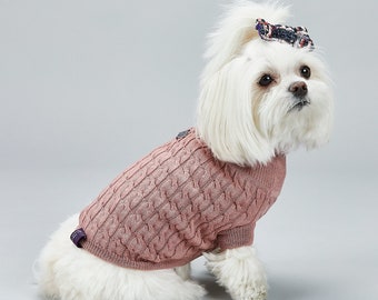 May include: A small white dog wearing a pink cable knit sweater with a black button. The dog has a black and white bow in its hair.