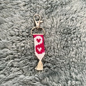 May include: A pink and white friendship bracelet keychain with a heart design and a tassel. The keychain is attached to a silver metal key ring.