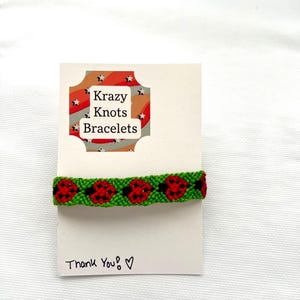 May include: A green friendship bracelet with red ladybug designs is displayed on a white card. The card has the text "Krazy Knots Bracelets" and "Thank You!" written on it. The bracelet is made of woven threads.