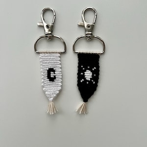 May include: Two handmade keychains with silver-tone clasps. One is white with a black letter "G", the other is black with a white sunburst design. Both have a woven design and a tassel.