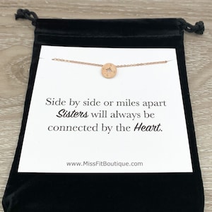 Puede incluir: Un collar de brújula de oro rosa exhibido en una tarjeta blanca dentro de una bolsa de terciopelo negro. La tarjeta dice: "Lado a lado o a millas de distancia, las hermanas siempre estarán conectadas por el corazón". El sitio web www.MissFitBoutique.com también está impreso.