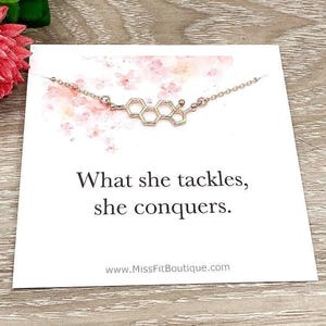 May include: A gold-coloured necklace featuring a molecular structure pendant, presented on a white card. The card displays the text "What she tackles, she conquers" and a floral design. The website address www.MissFitBoutique.com is also visible.