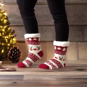 May include: Cozy red and white Christmas socks with a reindeer and tree pattern. The socks have a soft, white, faux fur trim at the top. The socks are worn with black leggings on a wooden floor.