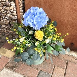 May include: A floral arrangement in a green pot features a large, light blue hydrangea, a yellow rose, and various greenery. The arrangement includes small white and yellow flowers, and eucalyptus leaves. The pot sits on a brick surface.