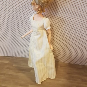 Puede incluir: Una muñeca blanca con el pelo rubio recogido en un moño alto, lleva un vestido blanco con rayas verticales. La muñeca está de pie sobre un suelo de madera.