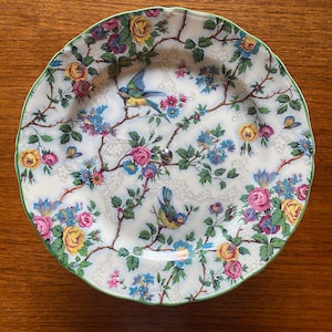 May include: A two-tiered serving plate with a floral and bird design. The plate features a white background with pink, yellow, and blue flowers, and two blue birds. The rim is green, and a silver handle connects the two tiers.