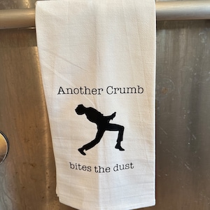 May include: White kitchen towel with black text that reads "Another Crumb bites the dust". A black silhouette of a person falling is in the center of the towel.