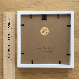 May include: A square white picture frame with a brown backing. The frame has black clips to hold the backing in place. A wooden ruler with the text "PENNY PATCH DESIGNS" is next to the frame. The backing has a sticker that says "handmade with Love".