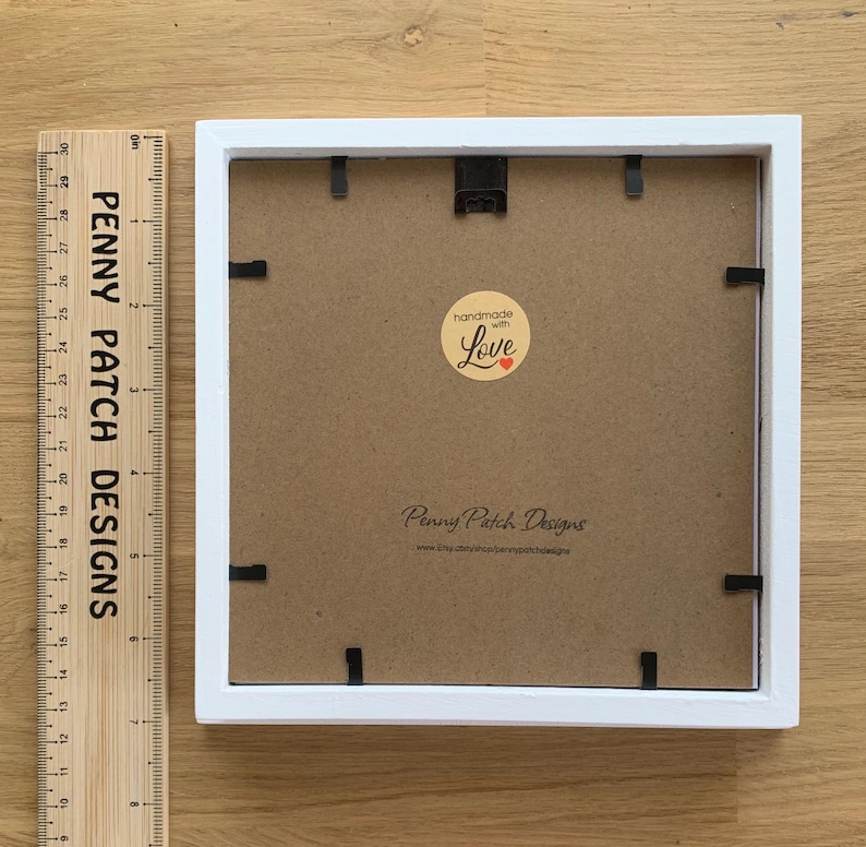 May include: A square white picture frame with a brown backing. The frame has black clips to hold the backing in place. A wooden ruler with the text "PENNY PATCH DESIGNS" is next to the frame. The backing has a sticker that says "handmade with Love".