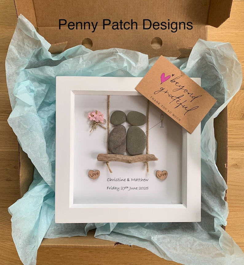 May include: A framed artwork featuring a couple on a swing, crafted from stones and driftwood. The white frame holds a scene with a small bouquet, two heart-shaped wooden accents with the word "Love", and a card that says "beyond grateful". The text "Penny Patch Designs" is visible.