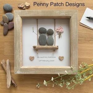 May include: A framed pebble art piece featuring two pebble figures on a swing, with a champagne glass and a bouquet. The frame is light brown, and the artwork includes the text "Mr and Mrs Rallison 15th February 2025" and "Penny Patch Designs".
