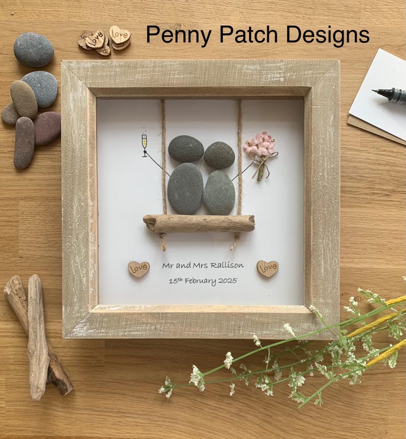 May include: A framed pebble art piece featuring two pebble figures on a swing, with a champagne glass and a bouquet. The frame is light brown, and the artwork includes the text "Mr and Mrs Rallison 15th February 2025" and "Penny Patch Designs".