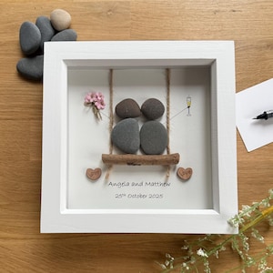 May include: A framed artwork featuring a swing made of stones. Two pairs of stones represent people on a swing, with a champagne glass and a small bouquet of pink flowers. The names "Angela and Matthew" and the date "25th October 2025" are written below.