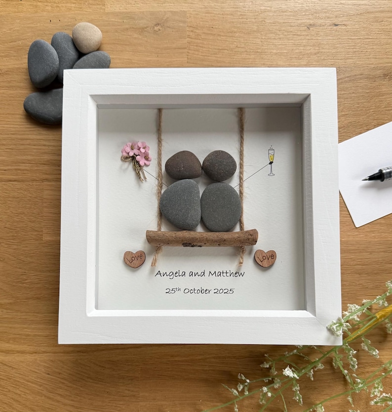 May include: A framed artwork featuring a swing made of stones. Two pairs of stones represent people on a swing, with a champagne glass and a small bouquet of pink flowers. The names "Angela and Matthew" and the date "25th October 2025" are written below.