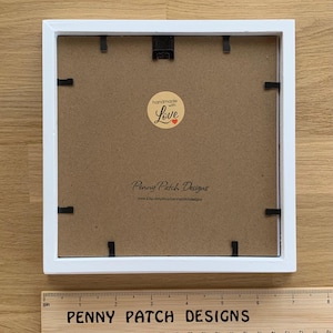 May include: A square white picture frame with a brown backing. The frame has black clips to hold the backing in place. A wooden ruler with the text "PENNY PATCH DESIGNS" is at the bottom. The backing has a sticker that says "handmade with Love".