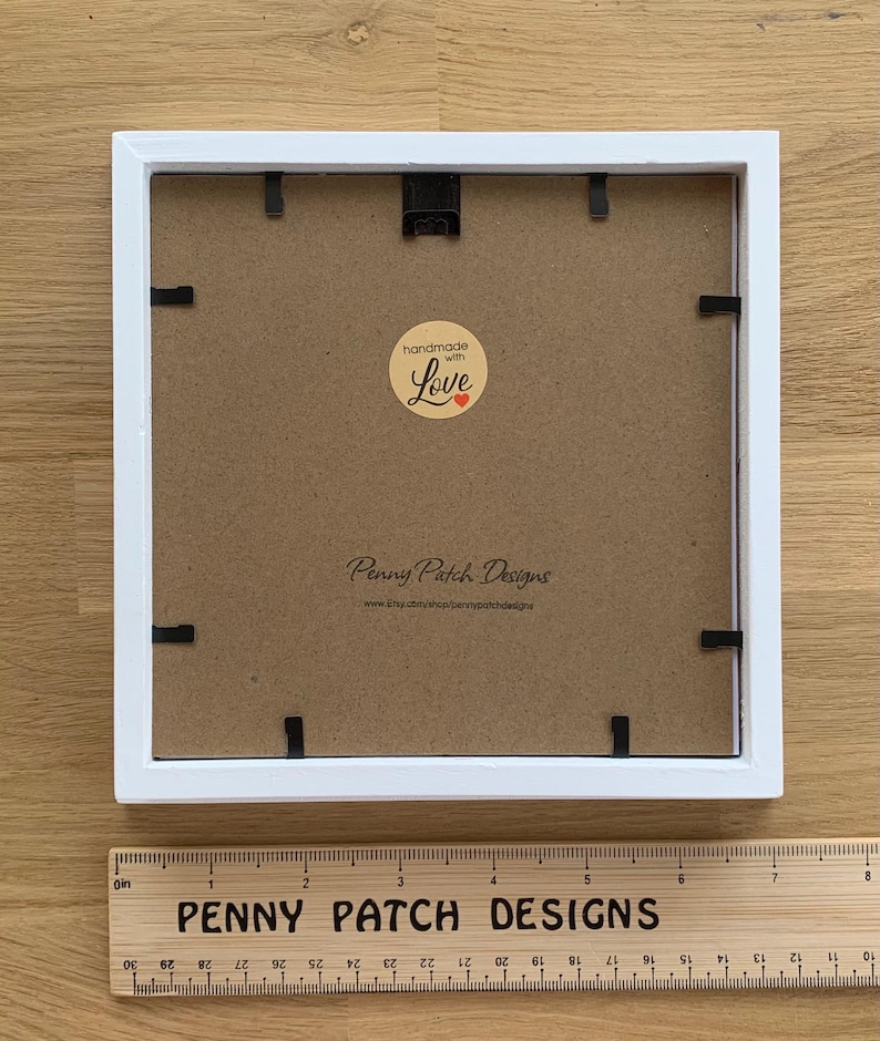 May include: A square white picture frame with a brown backing. The frame has black clips to hold the backing in place. A wooden ruler with the text "PENNY PATCH DESIGNS" is at the bottom. The backing has a sticker that says "handmade with Love".