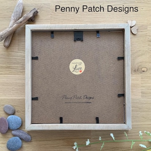 May include: A square, light-colored wooden picture frame with a brown backing. The frame has a black hanging hook and several black clips. The text "Penny Patch Designs" is visible, along with a "handmade with Love" sticker. Decorative stones and driftwood are nearby.