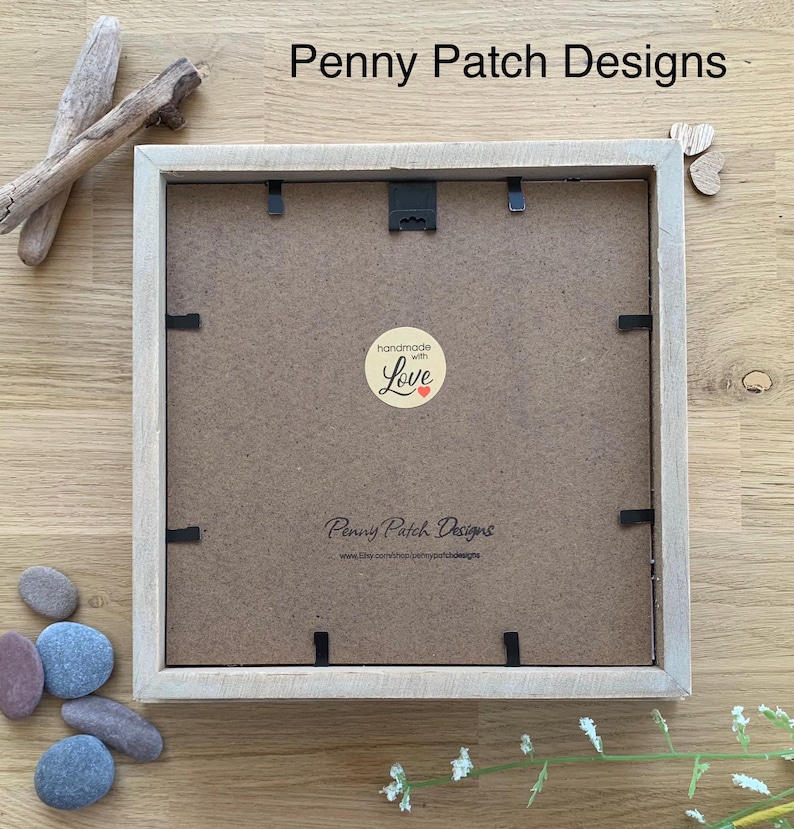 May include: A square, light-colored wooden picture frame with a brown backing. The frame has a black hanging hook and several black clips. The text "Penny Patch Designs" is visible, along with a "handmade with Love" sticker. Decorative stones and driftwood are nearby.