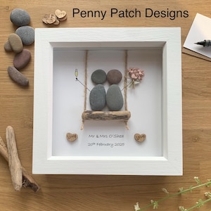 May include: A framed pebble art piece with a white frame. The art depicts two pebble figures on a swing, with a champagne glass and flowers. The text "Mr & Mrs O'Shea 20th February 2025" is below the swing. The words "Penny Patch Designs" are at the top.