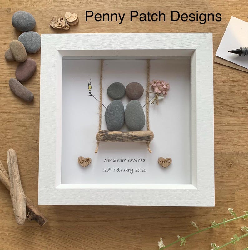 May include: A framed pebble art piece with a white frame. The art depicts two pebble figures on a swing, with a champagne glass and flowers. The text "Mr & Mrs O'Shea 20th February 2025" is below the swing. The words "Penny Patch Designs" are at the top.