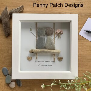 May include: A framed pebble art piece featuring two pebble figures on a driftwood swing. The artwork includes a champagne glass, a bouquet of flowers, and the names "Verena and Francesco" with the date "4th October 2024".