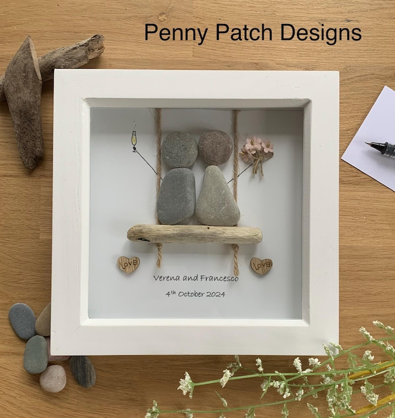May include: A framed pebble art piece featuring two pebble figures on a driftwood swing. The artwork includes a champagne glass, a bouquet of flowers, and the names "Verena and Francesco" with the date "4th October 2024".