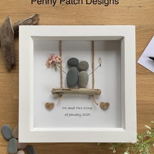 May include: White framed artwork featuring a pebble art scene of a couple on a swing. The swing is made of driftwood and rope, with a small bouquet of pink flowers. The artwork includes the text "Mr and Mrs King 18 January 2025".